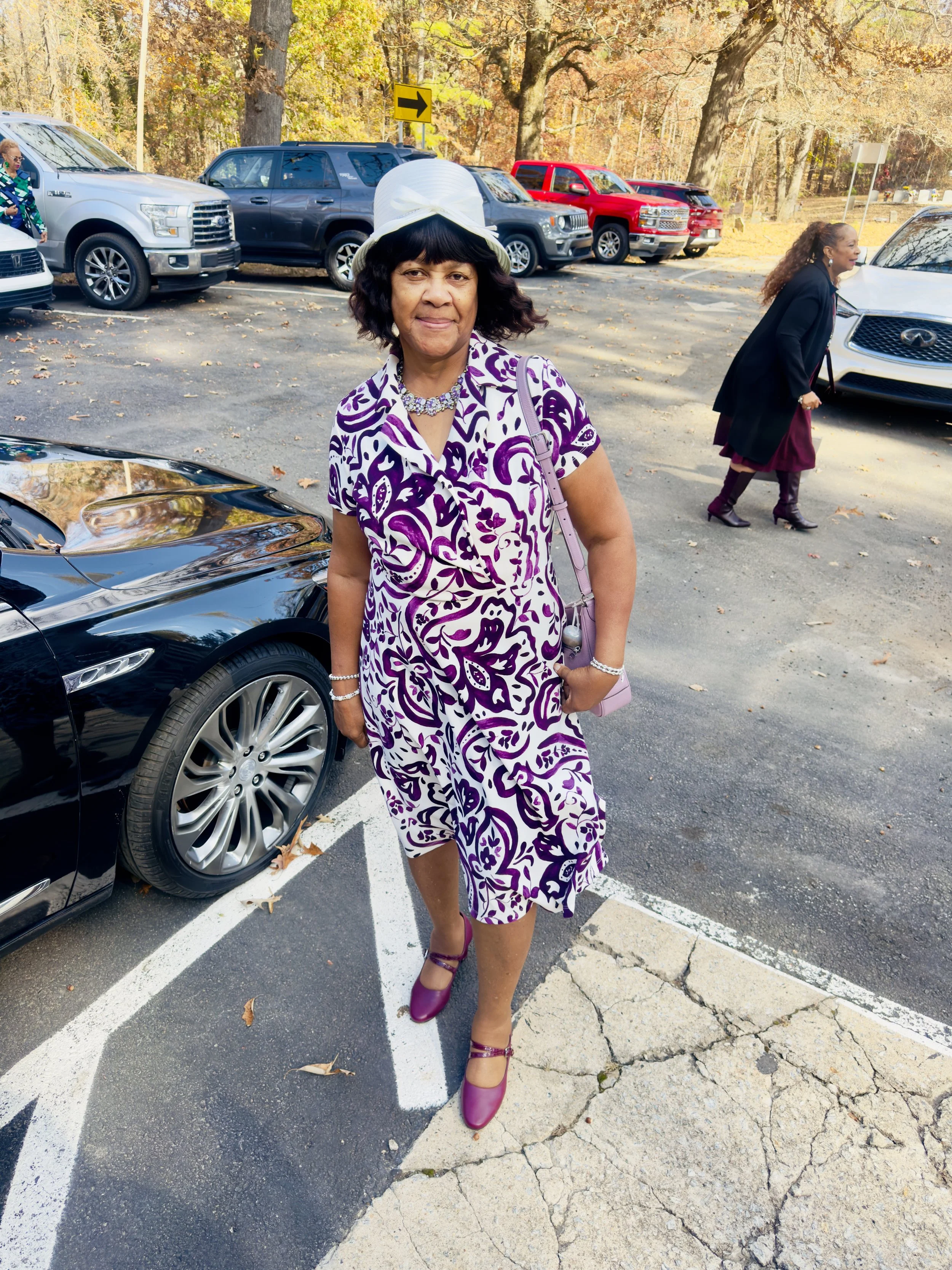 A woman in a purple and white patterned dress with purple shoes, wearing a white hat, standing in a parking lot with parked cars and trees with fall foliage in the background.