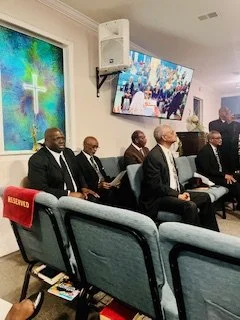 Group of men dressed in formal suits sitting on church pews during a service or event, with a large screen displaying a photo of a crowd in the background.