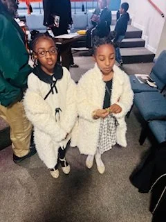 Two young girls standing side by side at an indoor event, both wearing fluffy white jackets and white dresses, with others in the background.