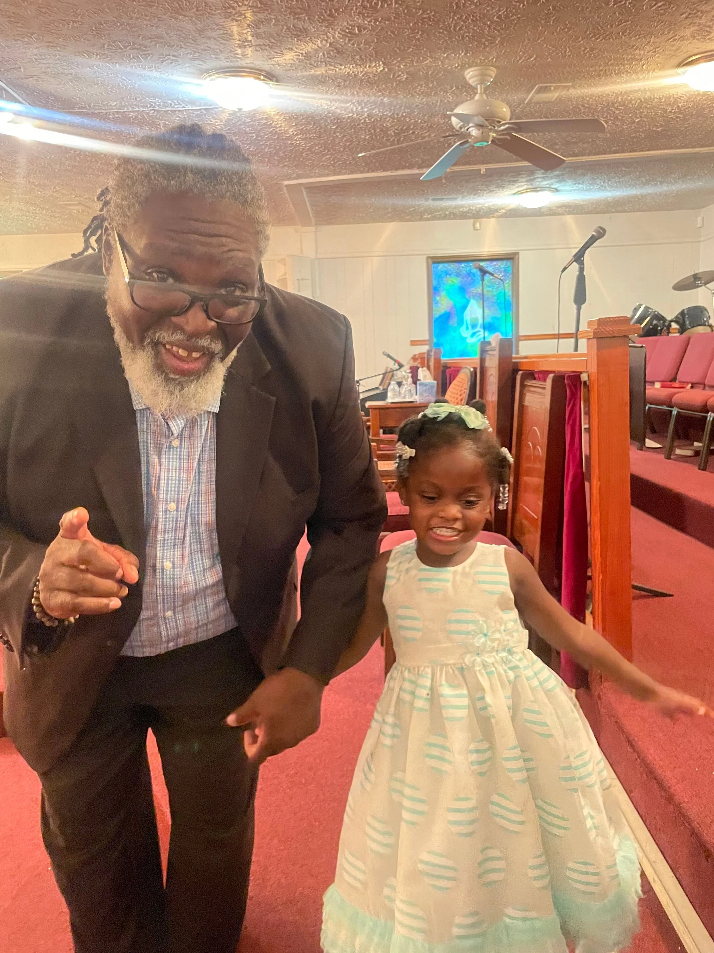 An older man with glasses and a beard, dressed in a suit, leaning forward and smiling, pointing towards the camera, standing next to a young girl in a white dress with light blue stripes, in a church setting with pews, microphones, and a stained glas