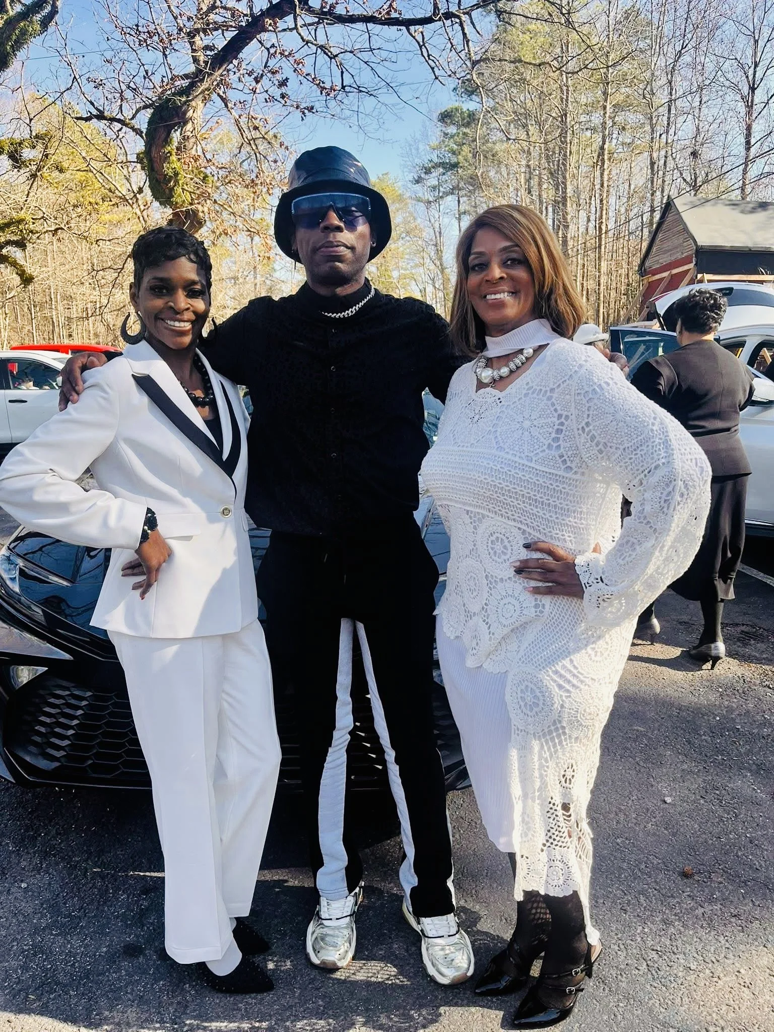 Three people standing outdoors in front of parked cars and trees, dressed in stylish outfits, smiling at the camera.