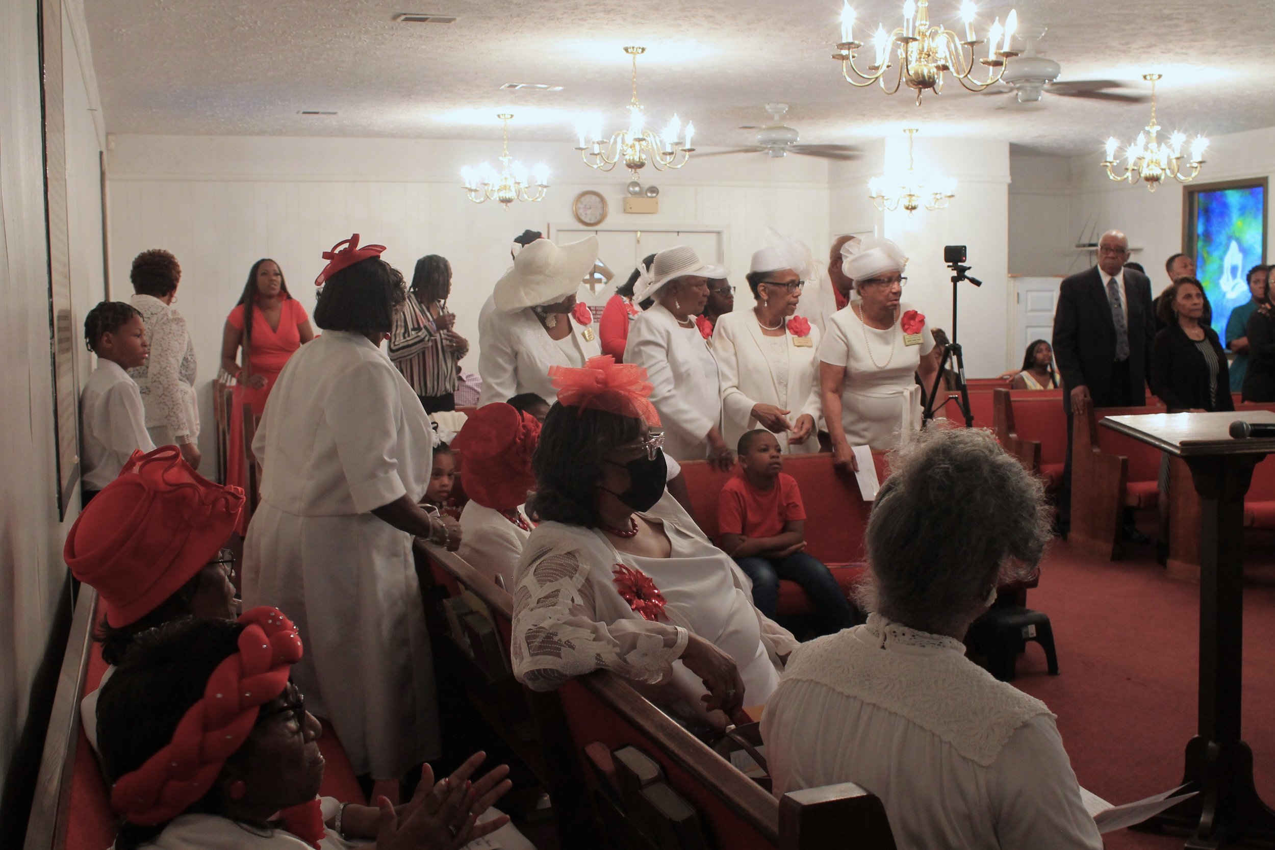 A gathering of African American women and children in a church, dressed in white and red attire, some wearing hats or head coverings, participating in a ceremony or service. The church interior is decorated with chandeliers and a large screen on the 