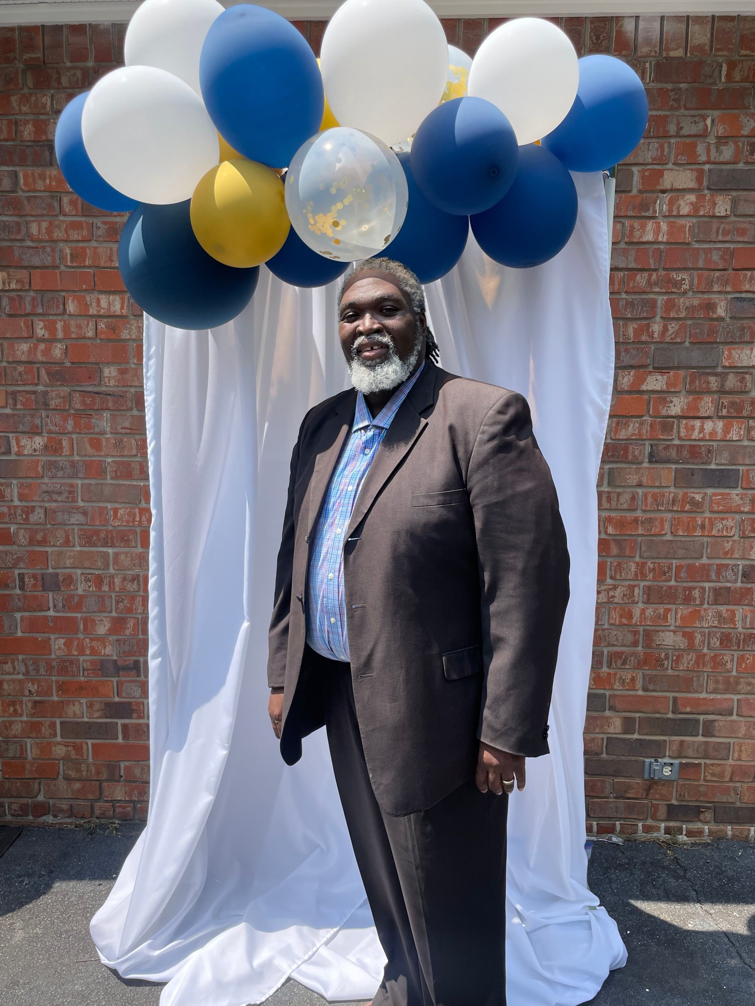 A man with gray hair and beard, wearing a brown suit and plaid shirt, standing in front of a white fabric backdrop with blue, white, yellow, and clear balloons. In front of a brick wall.