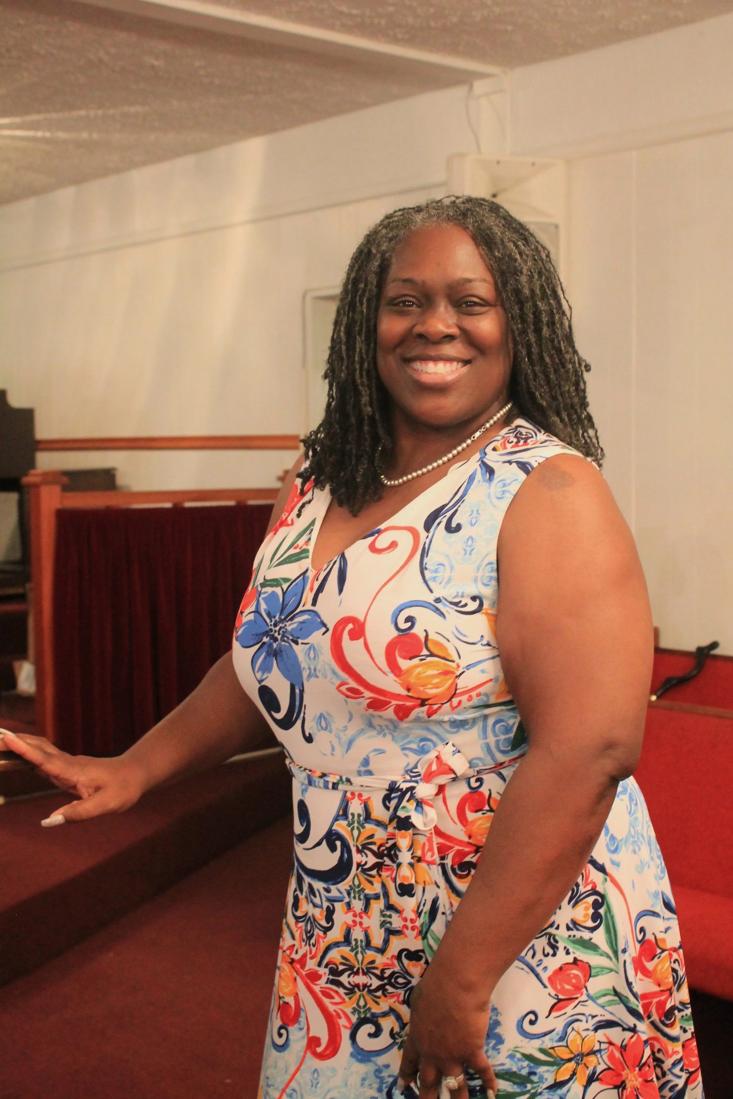 A smiling woman in a colorful floral dress standing inside a room with wooden furniture and red cushioned seating.