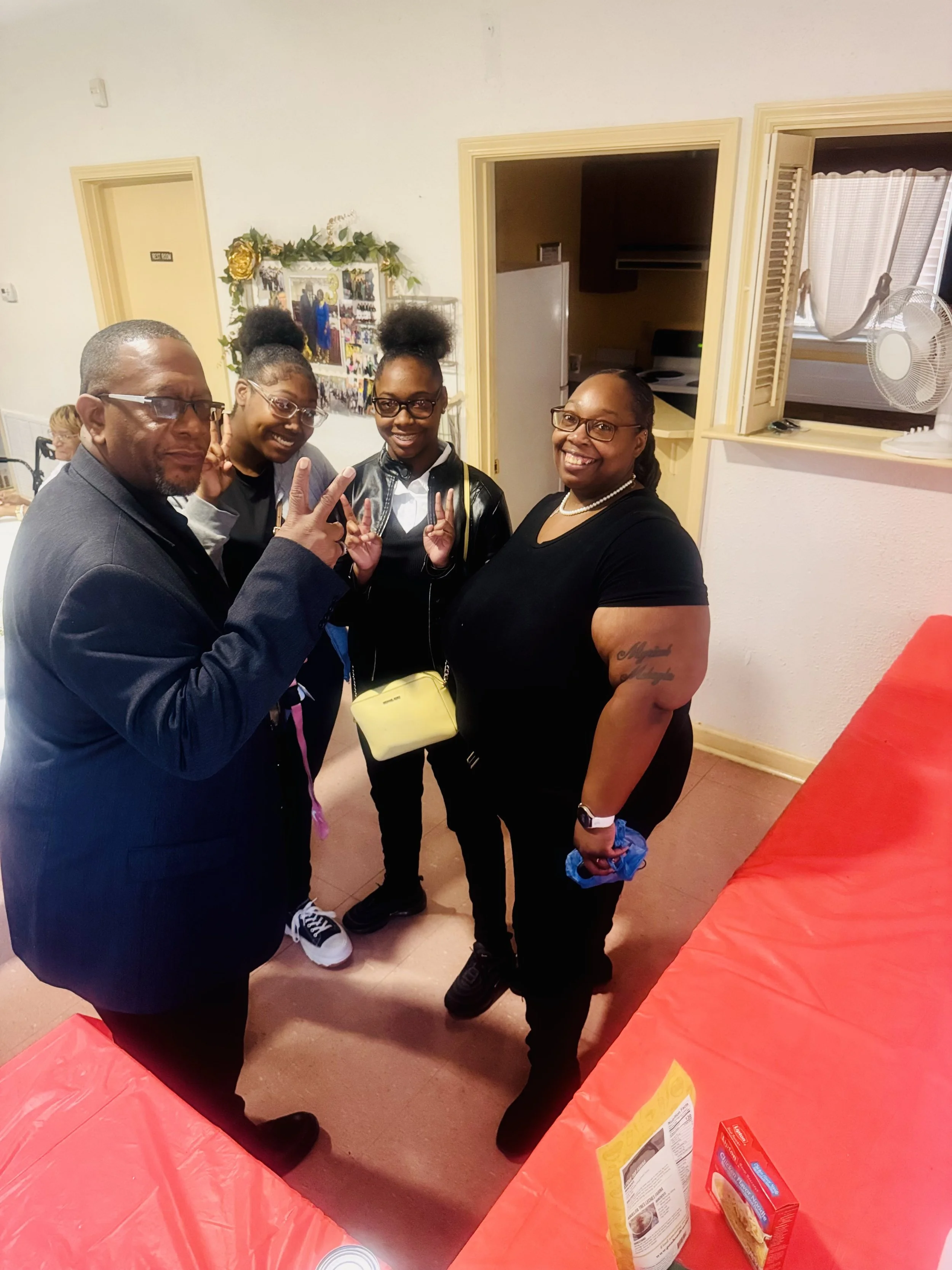 Four people standing together indoors, smiling at the camera, with one making a peace sign. The group includes one man with glasses and three women, one of whom is wearing glasses. They appear to be at a social gathering or party.