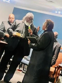 Three people at an indoor event, with one person holding a microphone and a tray, engaging in conversation.