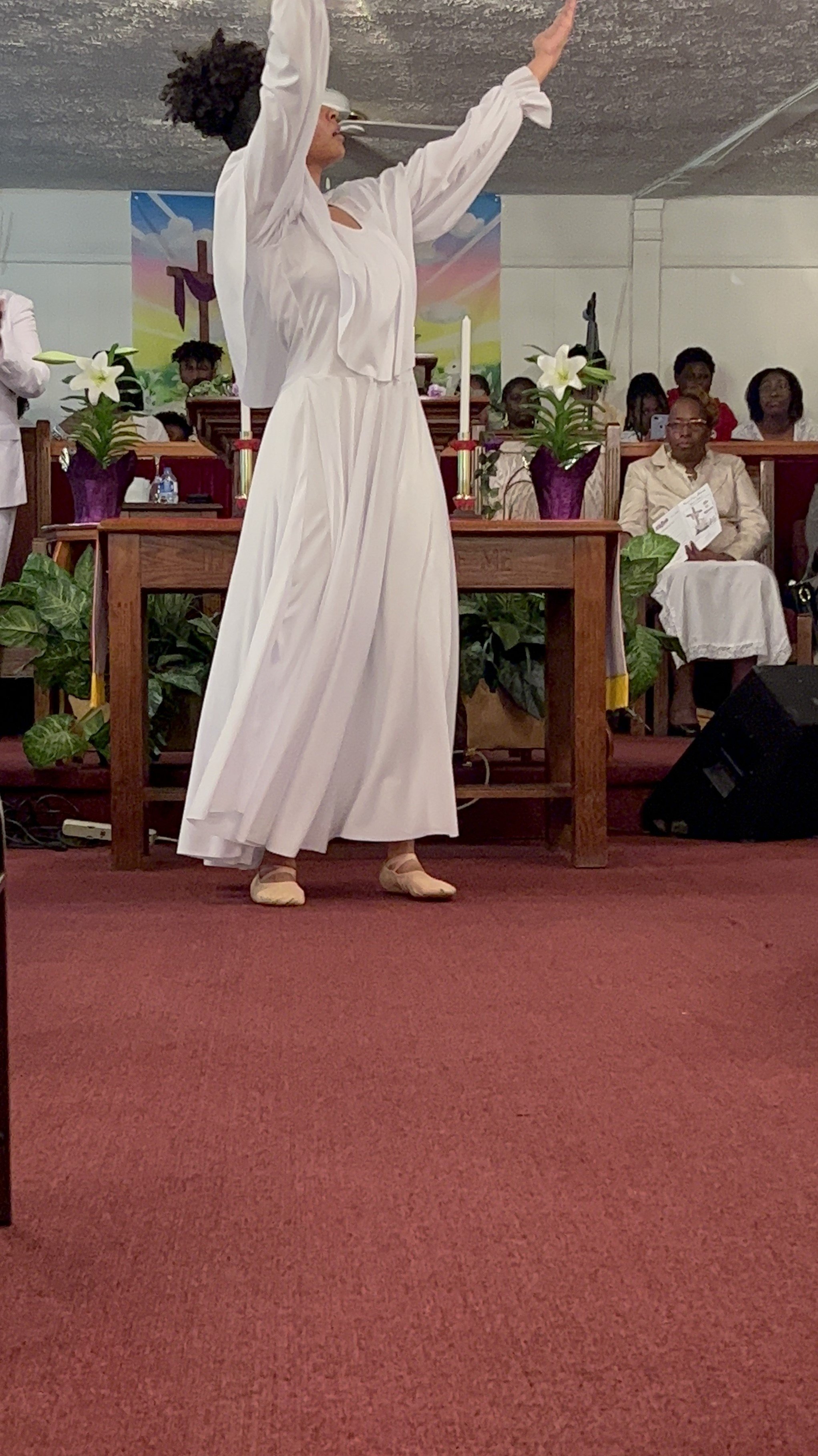 A woman in a white dress and ballet shoes stands with arms extended during a service or performance in a church, with worshippers seated behind her.