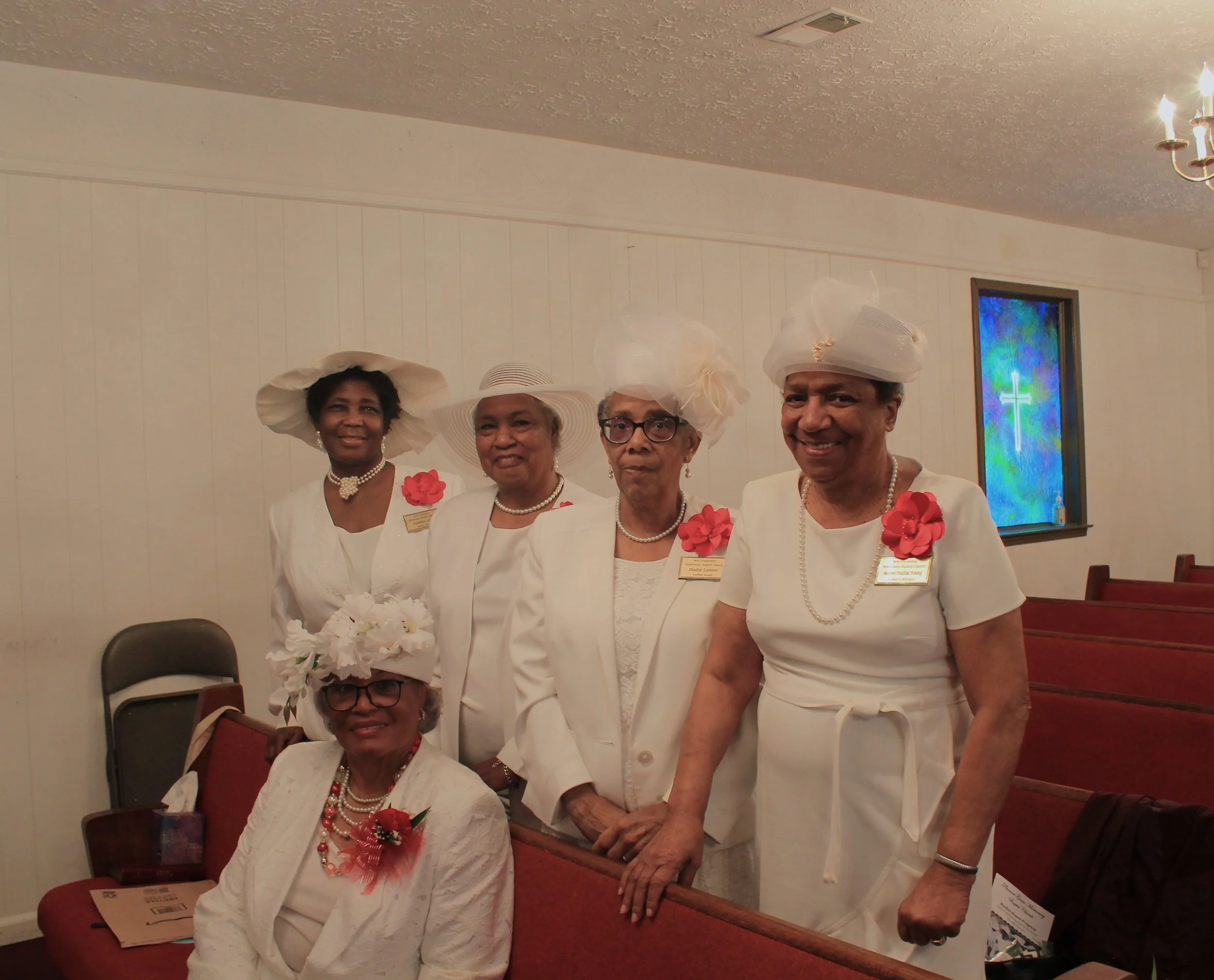 Five women dressed in white, some wearing white hats with decorative flowers, standing and sitting inside a church. They are wearing name tags and some have red or white flowers pinned to their clothing. The background includes wooden pews and staine