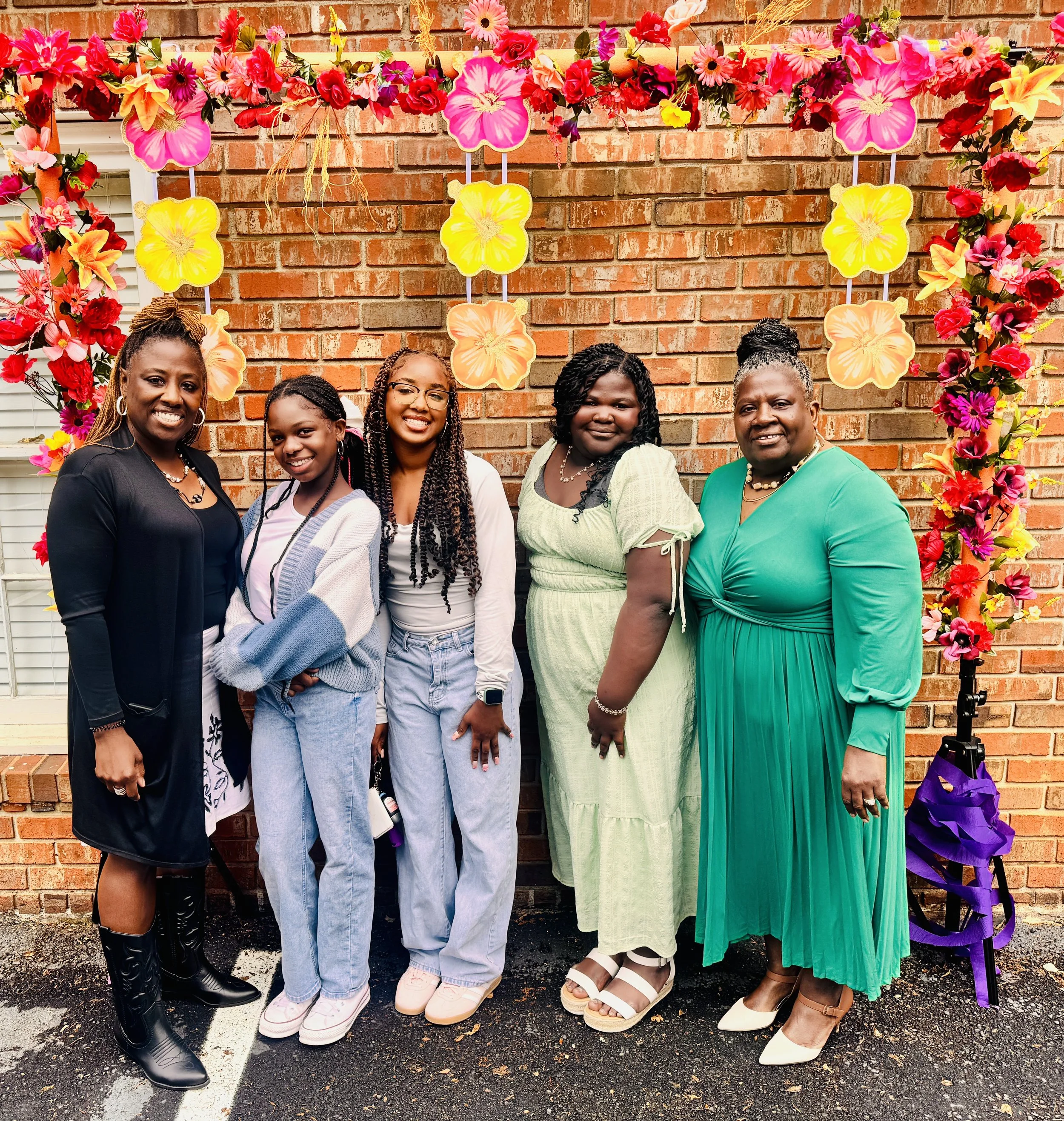 Five women standing in front of a brick wall decorated with pink, yellow, red, and purple floral arrangements for a celebration or event.