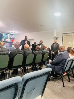 A group of people attending a meeting in a conference room, with some seated and others standing at the front, where a speaker is addressing the audience.