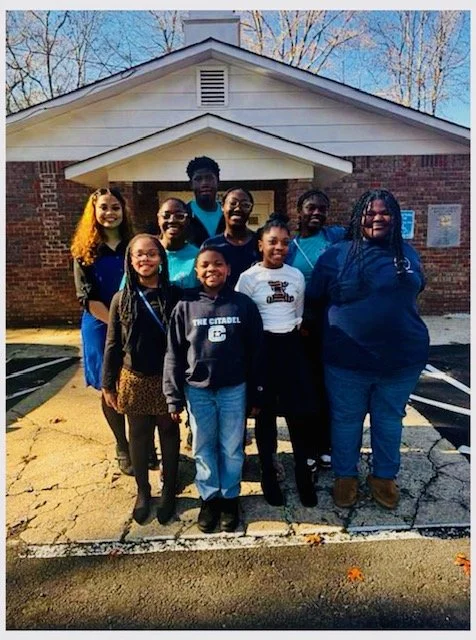 Group of nine children and teenagers standing together outside a brick building, smiling.