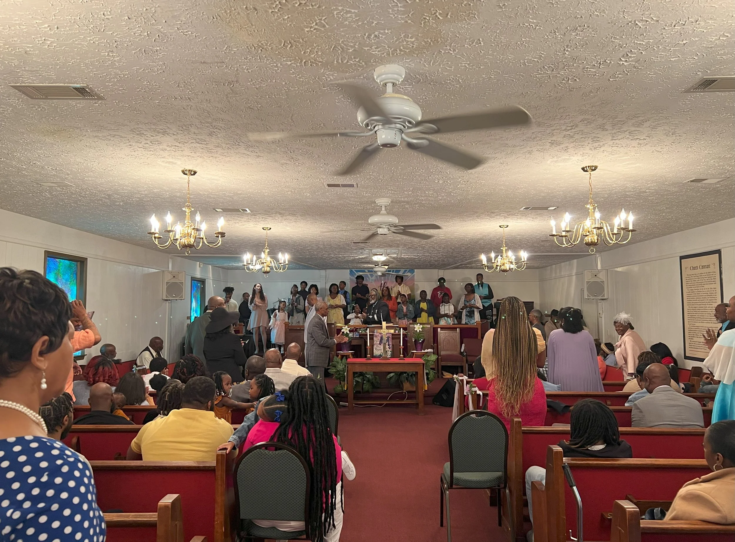 A church congregation gathered for a service or celebration, with people standing, sitting, and singing, decorated with chandeliers and framed artwork.