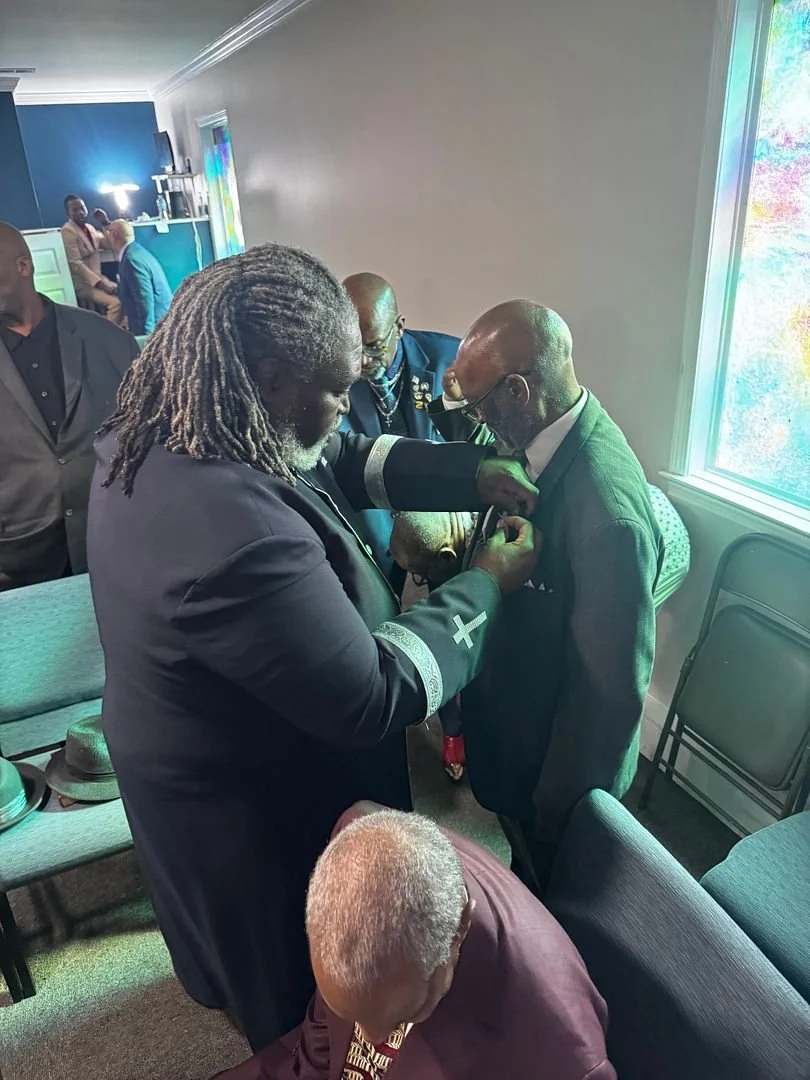 A man in a black suit with a white cross on the sleeve is receiving a pin or badge from another man in a suit, while a woman with gray dreadlocks stands beside them and an elderly person with white hair is seated in front. Several other people are vi