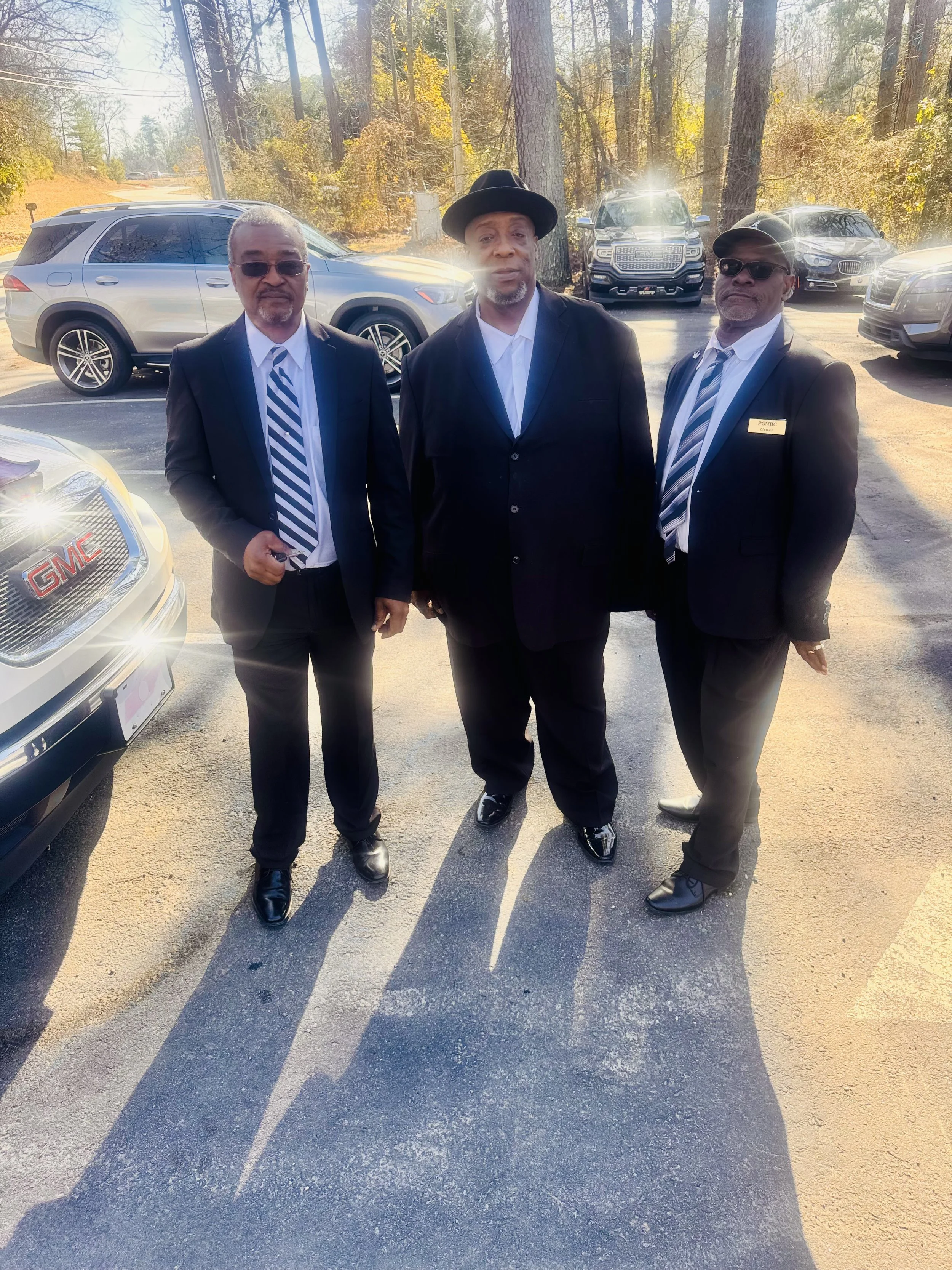 Three men dressed in suits standing in a parking lot surrounded by parked cars and trees.