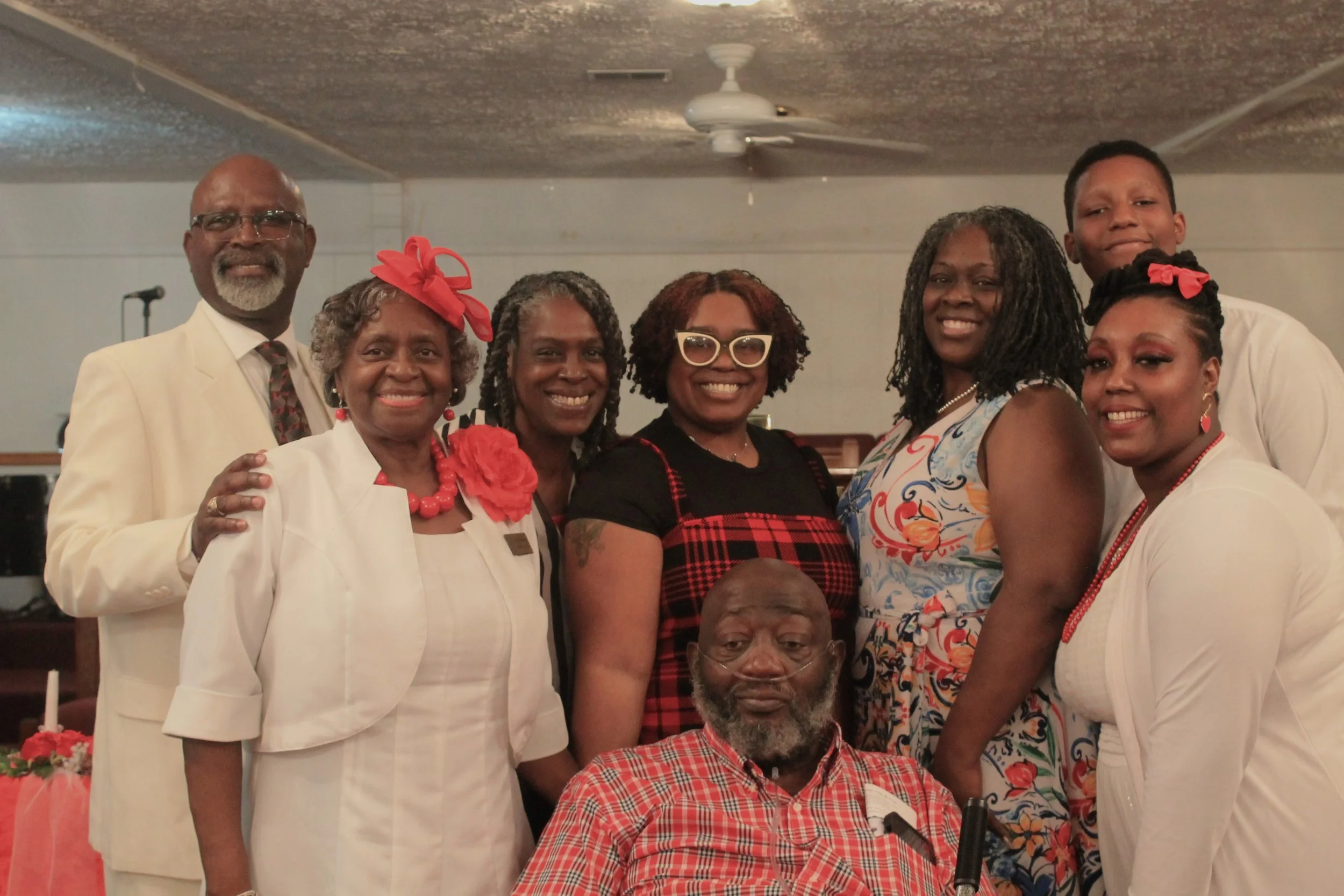 A group of eight African American people gathered in a room, dressed in formal and colorful attire, smiling for a photo. An elderly man with a gray beard is seated in front, and the others stand around him.