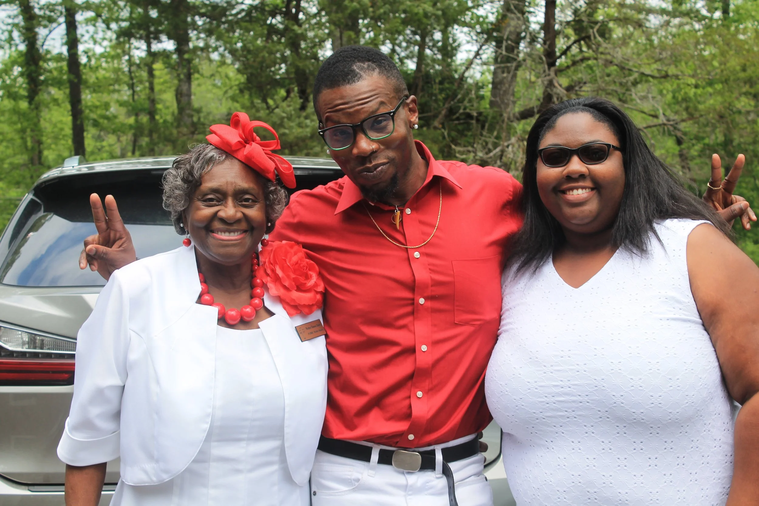 Three people standing outdoors in front of a car, smiling, two women and one man, making peace signs with their hands.