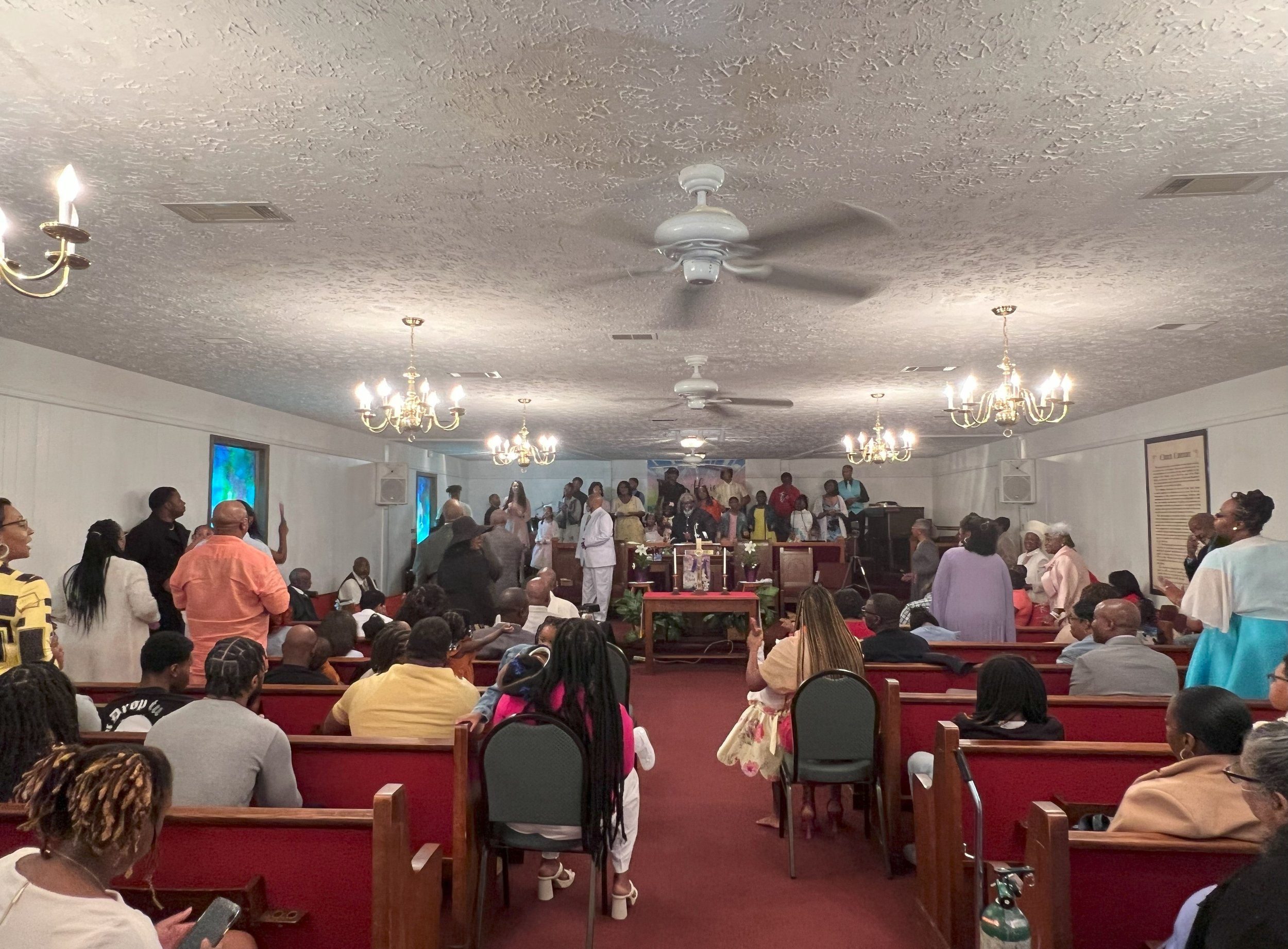Interior of a church filled with people, some standing and others seated in pews, attending a service or event with a stage area at the front, decorated with flowers and religious symbols, illuminated by hanging chandeliers.