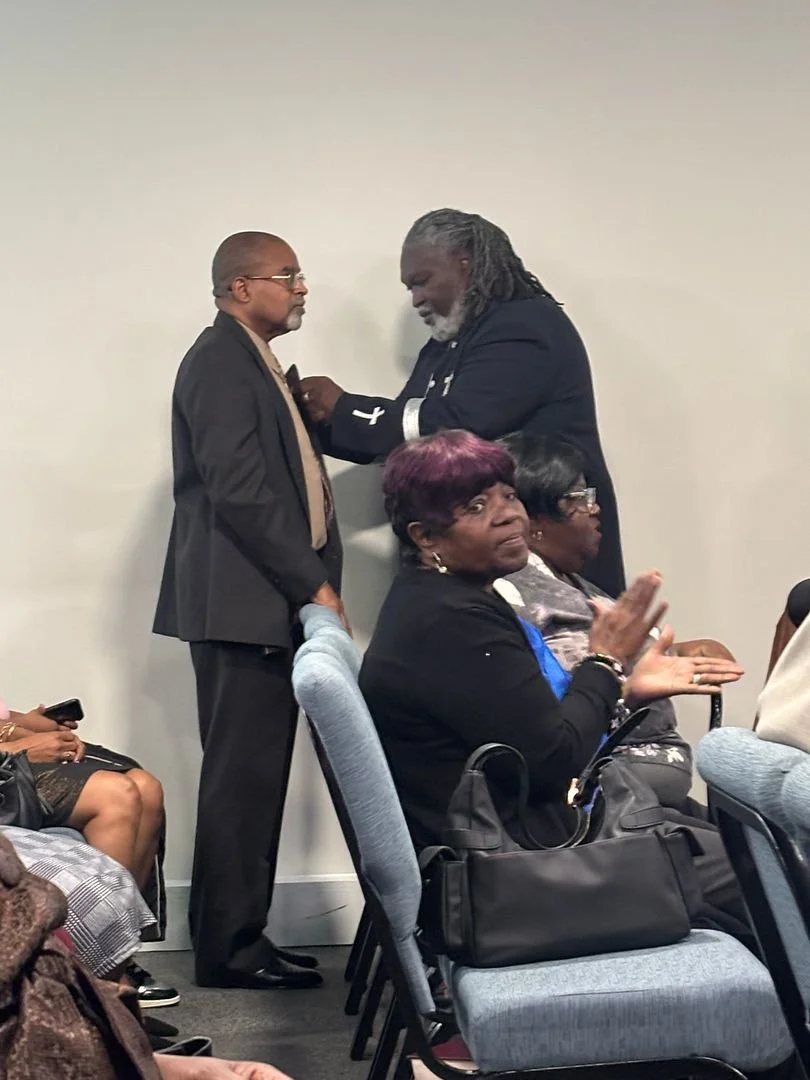Two men in suits share an intimate moment, with one placing a hand on the other's chest, while three women seated nearby look on, with one clapping.