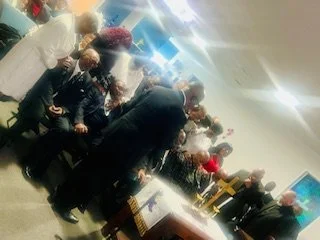 A group of people gathered around a table in a room, possibly a formal or religious event, with some dressed in dark clothing and headscarves, and others in casual attire.