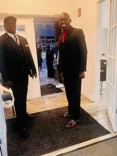 Two men standing inside a house near the entryway, dressed in formal suits, smiling at the camera.