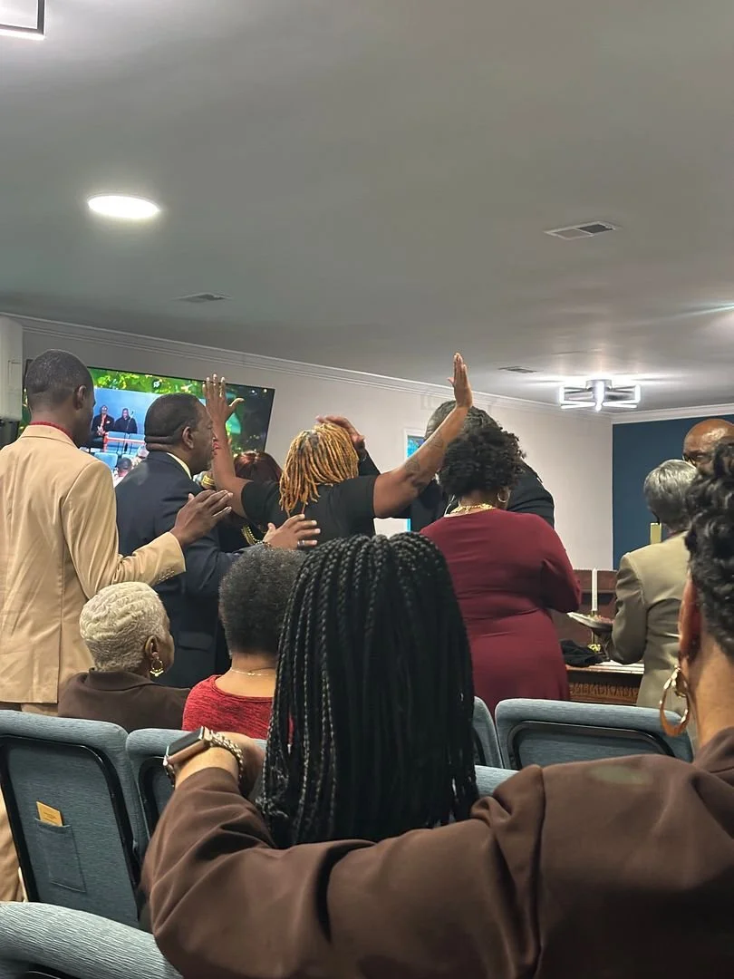 Group of people gathered indoors, some standing and raising hands, others seated, engaged in a religious or spiritual service.