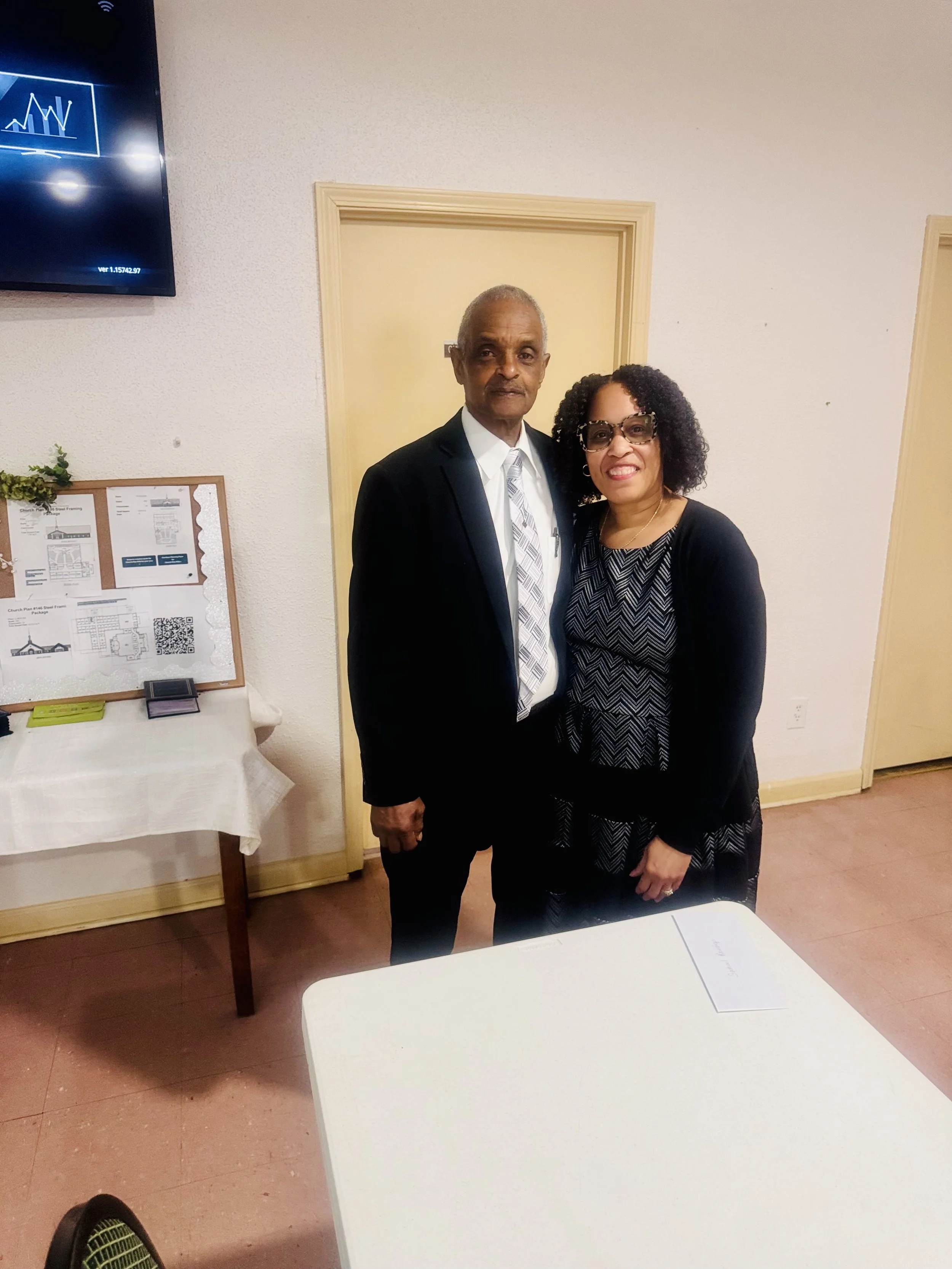 An elderly man and woman are standing together indoors, smiling at the camera. The man is wearing a dark suit with a white shirt and patterned tie, and the woman is dressed in a black and gray patterned dress with a black cardigan, wearing sunglasses