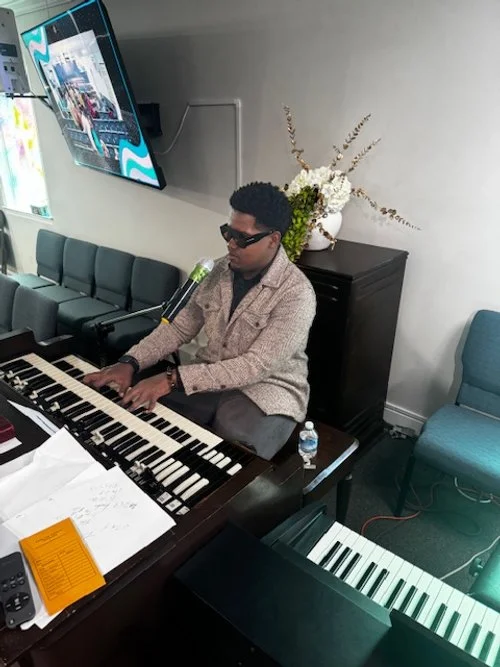 A man wearing sunglasses and a beige jacket playing an organ in a room with a floral arrangement, a blue chair, and a television mounted on the wall.