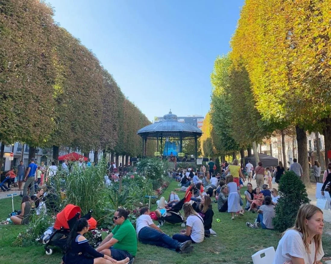 Un parc bondé avec des gens assis sur l'herbe et dans des chaises, une scène avec un gazebo au centre, entouré d'arbres aux feuilles automnales, un ciel clair.