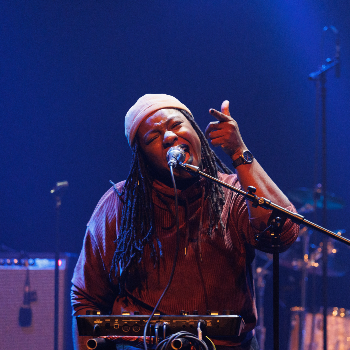 Une femme chante avec enthousiasme lors d'un concert, portant un pull rouge, un bandeau blanc et des dreadlocks, avec un synthétiseur devant elle.