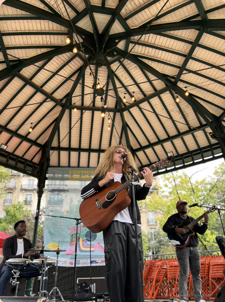 Un groupe de musiciens joue sur scène lors d'un festival en plein air sous une grande structure en bois, avec des arbres en arrière-plan.