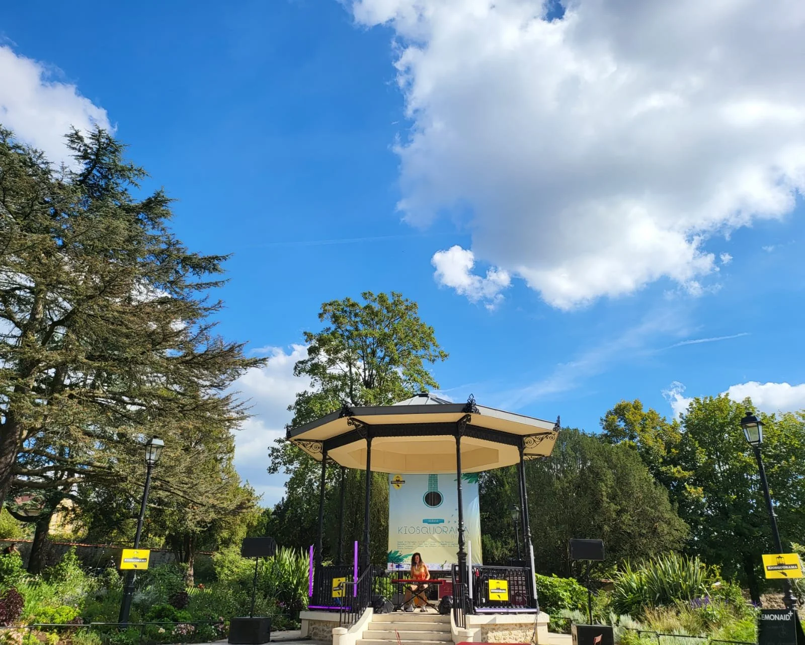 Une scène dans un parc avec un kiosque en forme de chapiteau, entourée d'arbres et de lampadaires, avec un panneau indiquant le nom 'Kiosquora' à l'arrière-plan.
