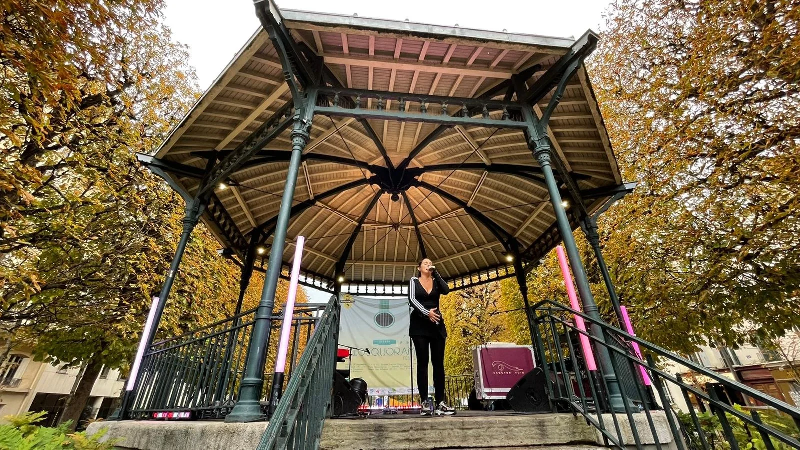 Une jeune femme chante en sanglant dans un micro sur une scène de kiosque en plein air, entourée d'arbres aux feuilles automnales.