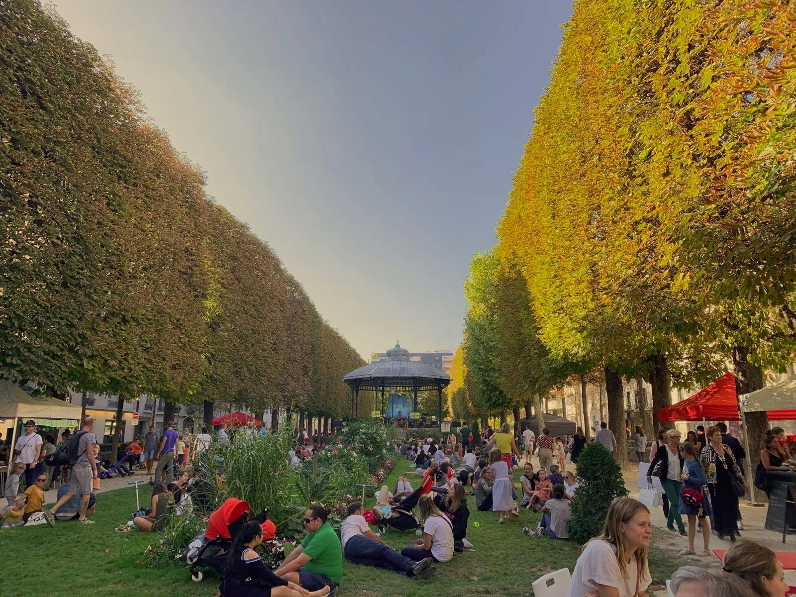Un parc public rempli de personnes assises ou marchant, entre des rangées d'arbres à feuilles vertes, avec une scène musique en arrière-plan en plein air, par un jour ensoleillé.