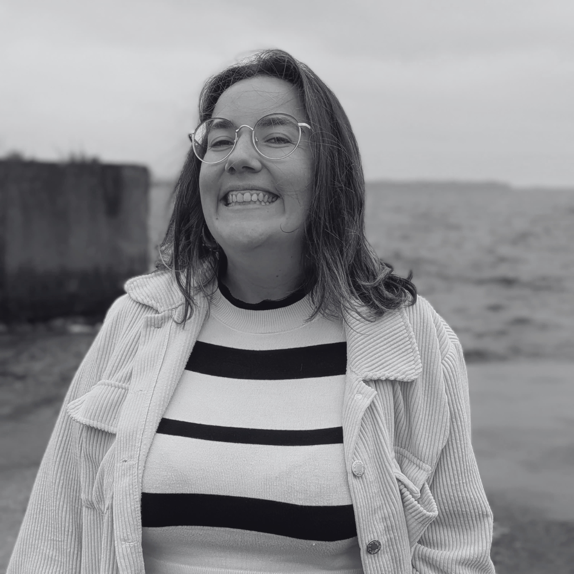 Jeune femme souriante portant des lunettes, un pull rayé et une veste en dehors, près de la mer.