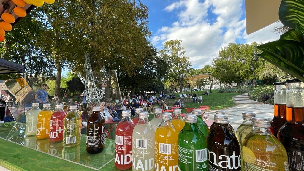Une rangée de bouteilles de différentes couleurs devant un public assis en plein air dans un parc, avec des arbres, un ciel bleu et des structures en métal en arrière-plan.
