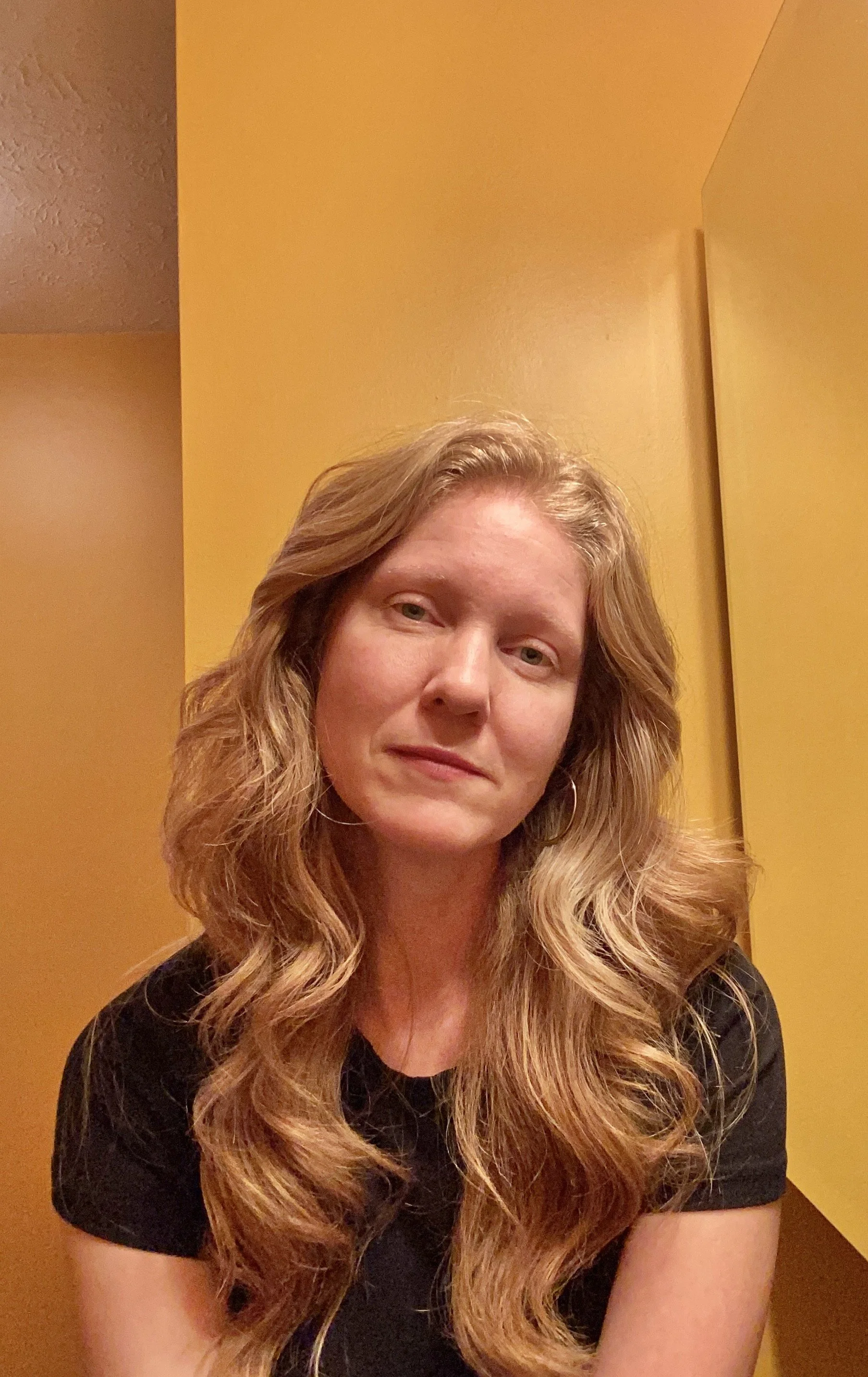 A woman with shoulder-length red hair, glasses, and hoop earrings smiling outdoors against a light gray background.