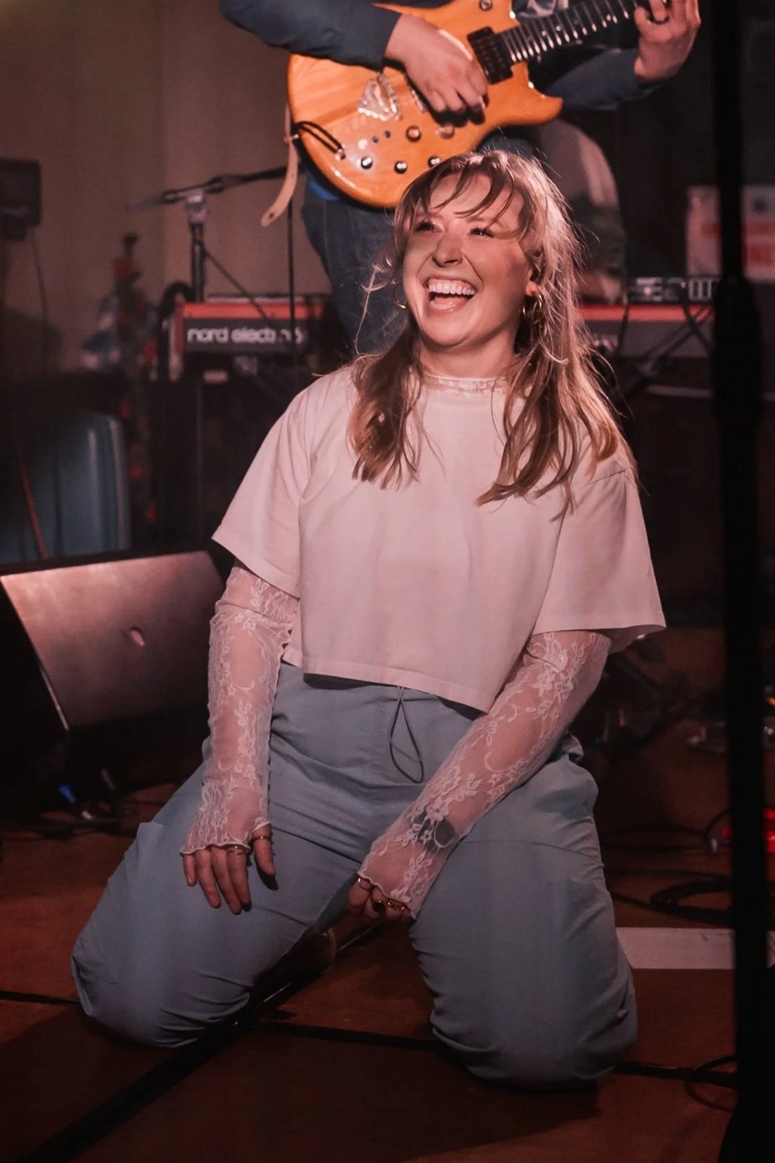 A young woman with long, curly blonde hair making a rock gesture with her hand, sticking out her tongue, in a vibrant, colorful music venue with purple, pink, and yellow lighting. A musician playing guitar is visible in the background.