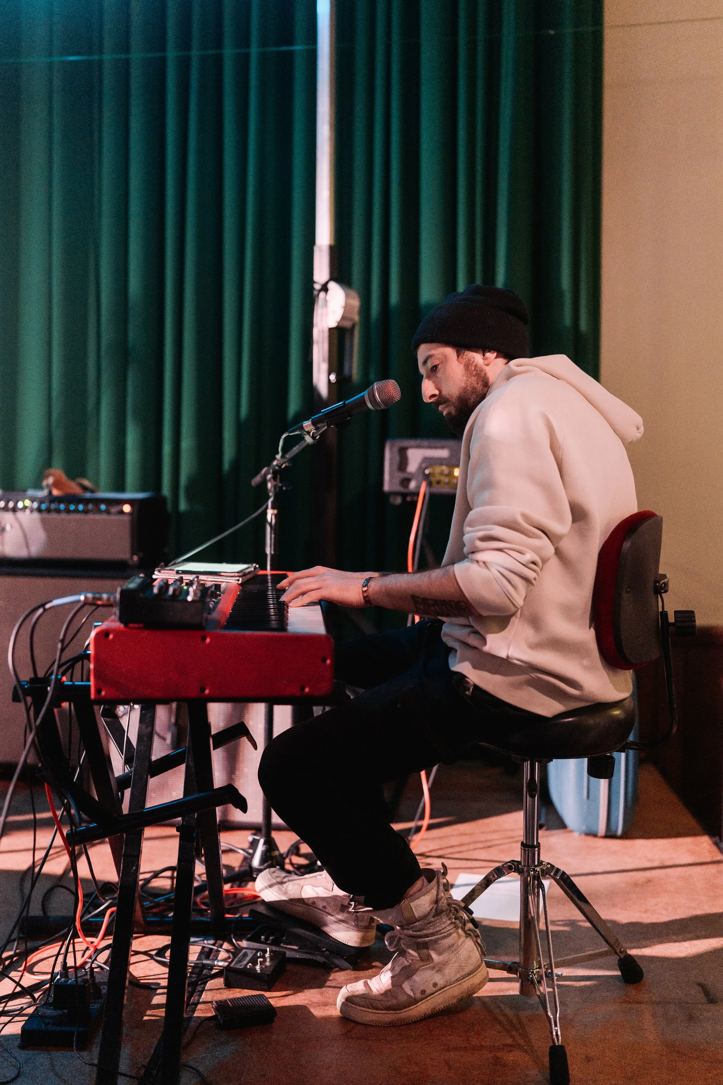 A young man in a hoodie and beanie playing a red Nord Electro 6D keyboard on stage, with a microphone in front of him, under purple and blue stage lighting.