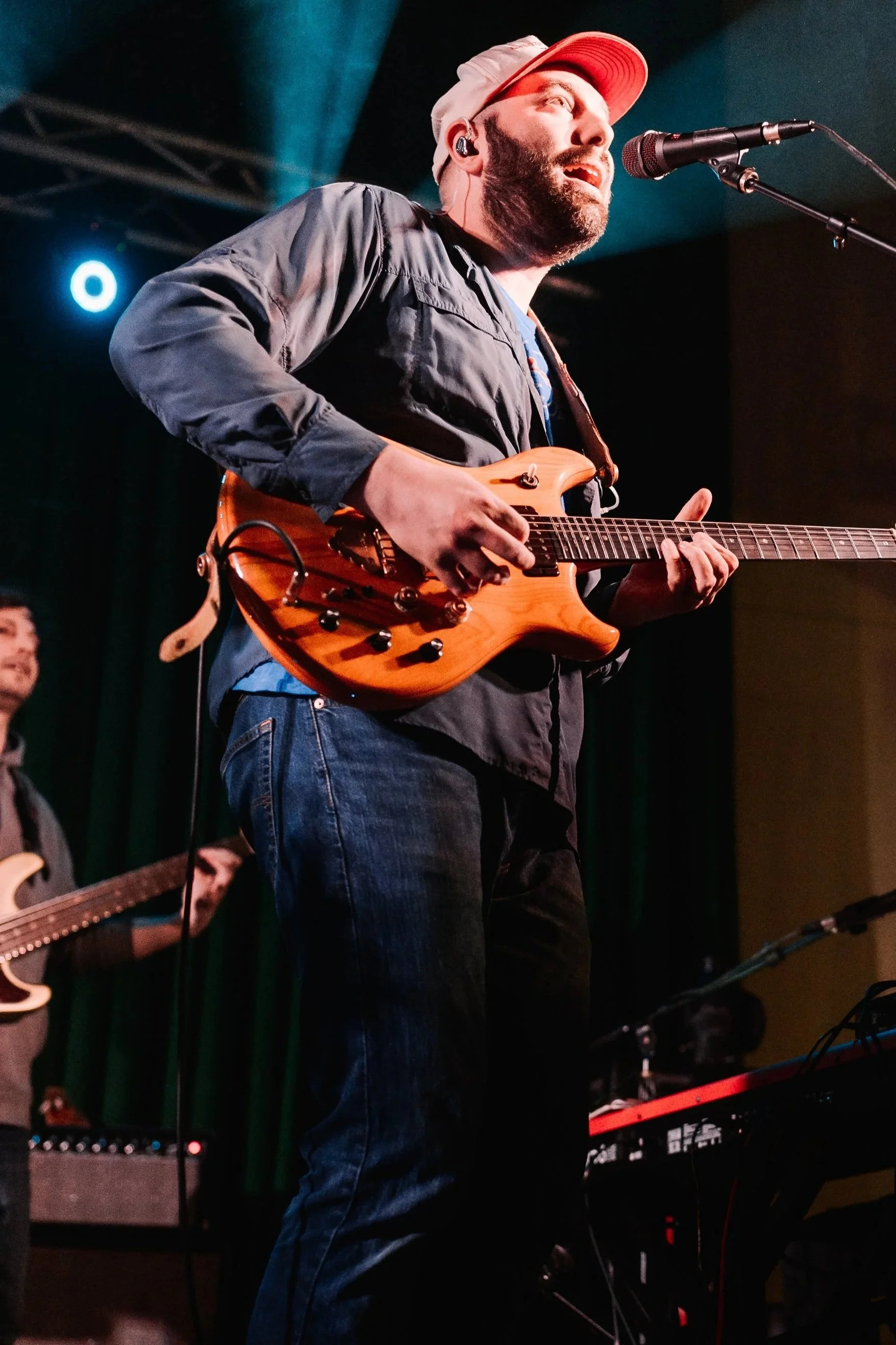 A man wearing sunglasses, a baseball cap, and a Hawaiian shirt playing an electric guitar on an outdoor stage.