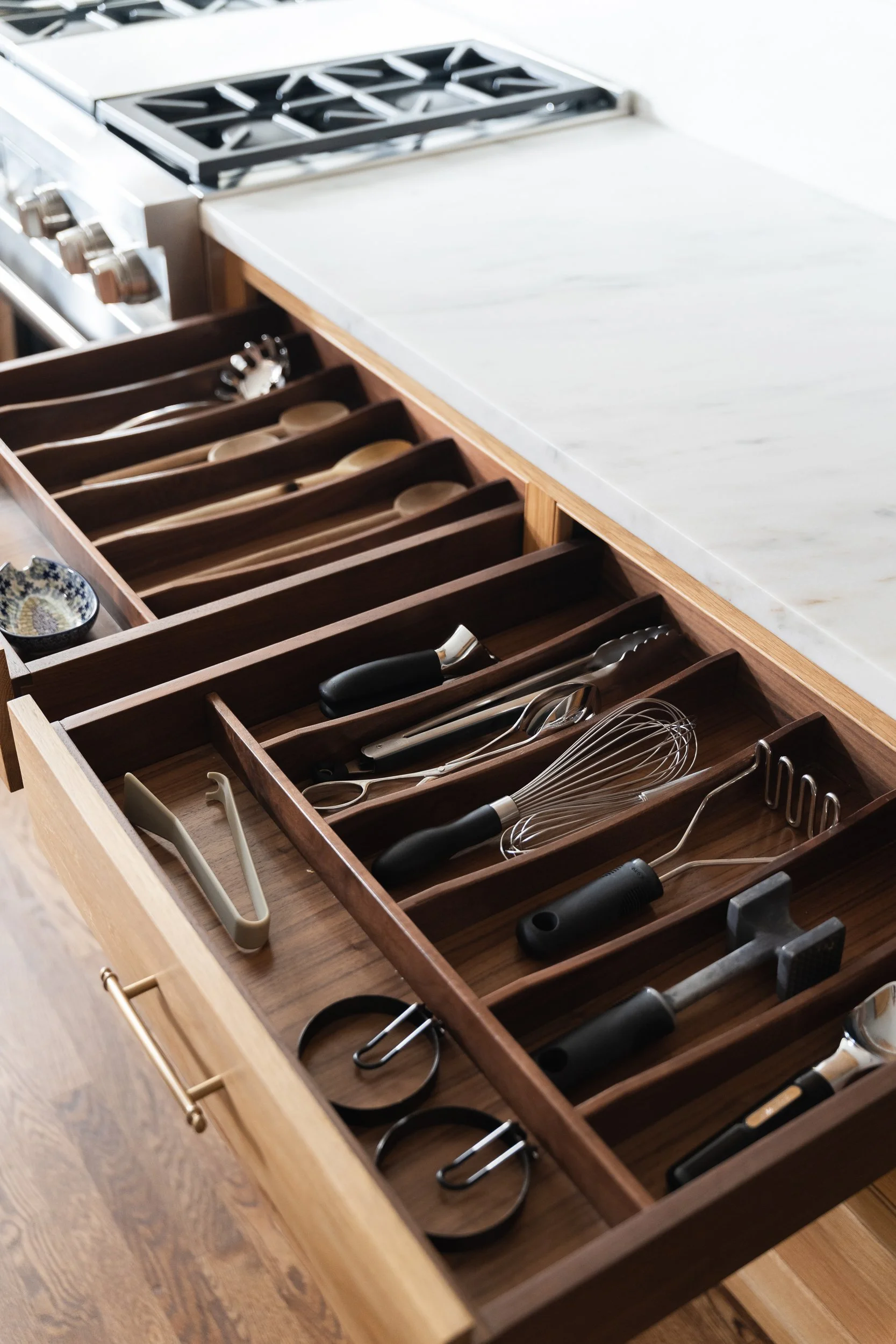 A wooden cutlery organizer with black-handled knives, spoons, and forks. The knives are arranged in a slot while the spoons and forks are in separate sections. A small black-handled pen is in a compartment on the left side.