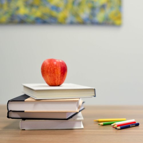 stack of books on a desk with apple on top; several colored pencils also on desk; classroom setting