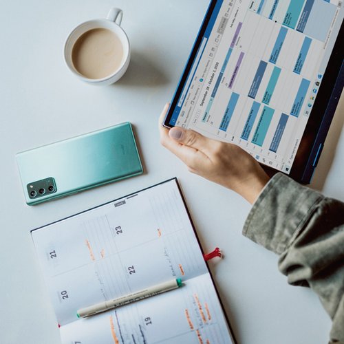 woman with digital and paper calendar at desk; blue phone and cup of coffee also on desk