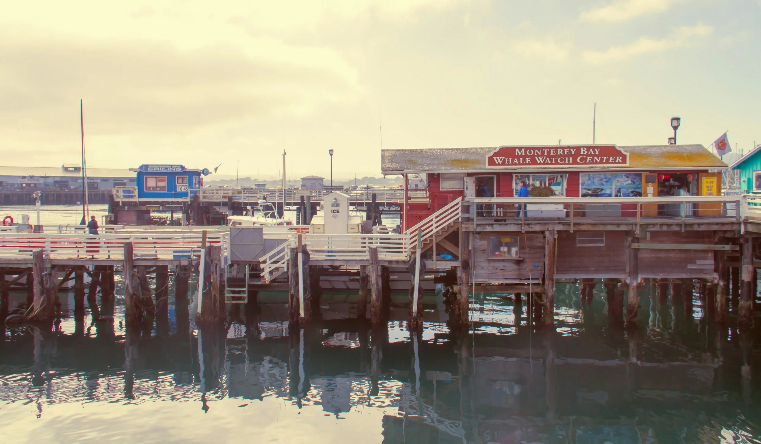 Wooden buildings line a pier on the Big Sur coast. One reads Monterey Bay Whale Watch Center. Drives & Detours Monterey Tour