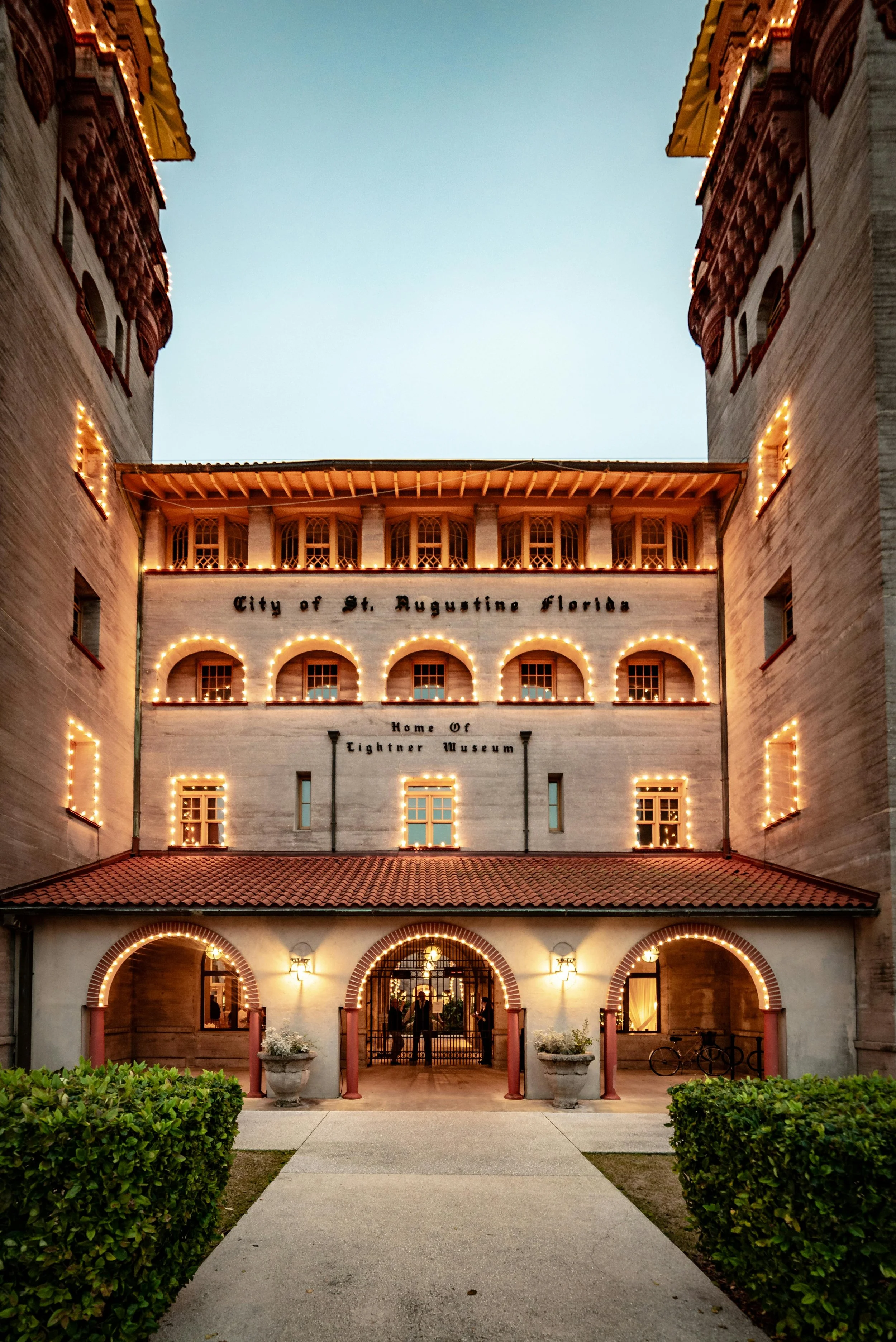The Lightner Museum is lit up at dusk. The shot is a close on of the entrabce and you can see the brickwork around the doors, the wooden windows and the arches that surround these