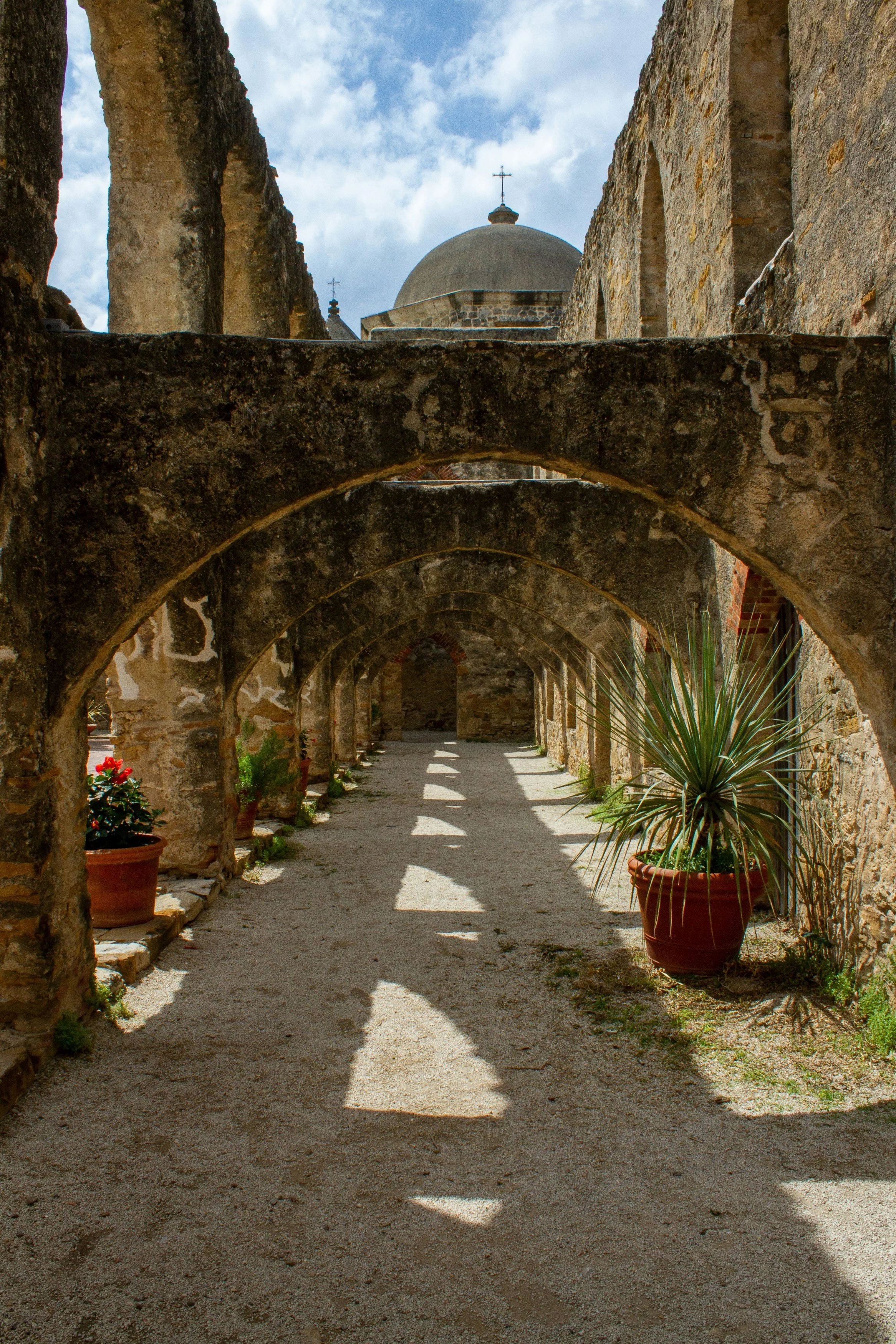 A cloister in Mission San José, San Antonio. It has no roof and the dome of the church can be seen at the end. The walkway is lined with plants