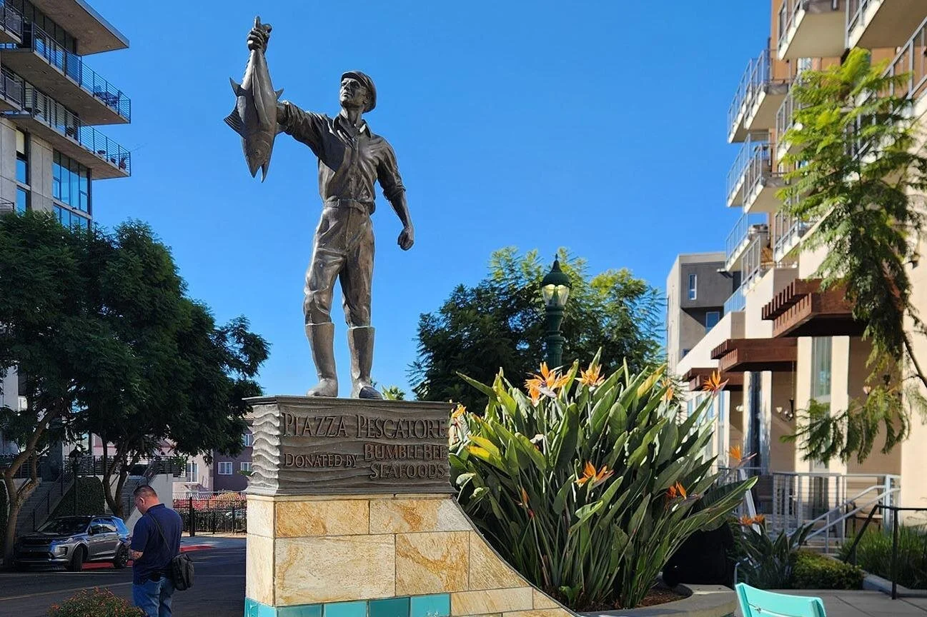 A statue of a man holding up a fish in celebration. The base of the statue reads: "Piazza Pescatore, Dontaed by Bumblebee Seafoods" The sky behind is a bright blue without any clouds