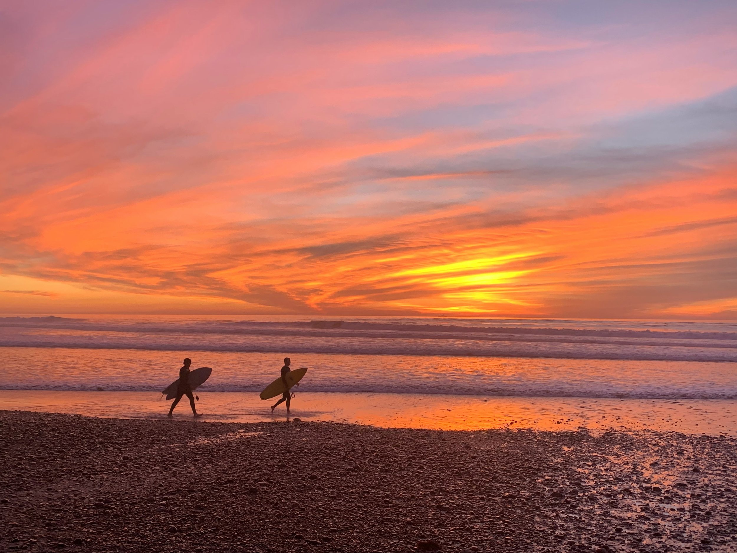 Two surfers walk towards the sea at sunset on Torrey Pines Beach, which you will see on your San Diego Driving Tour. The water is still and the sky is a deep orange, changed to bright yellows by cirrus clouds
