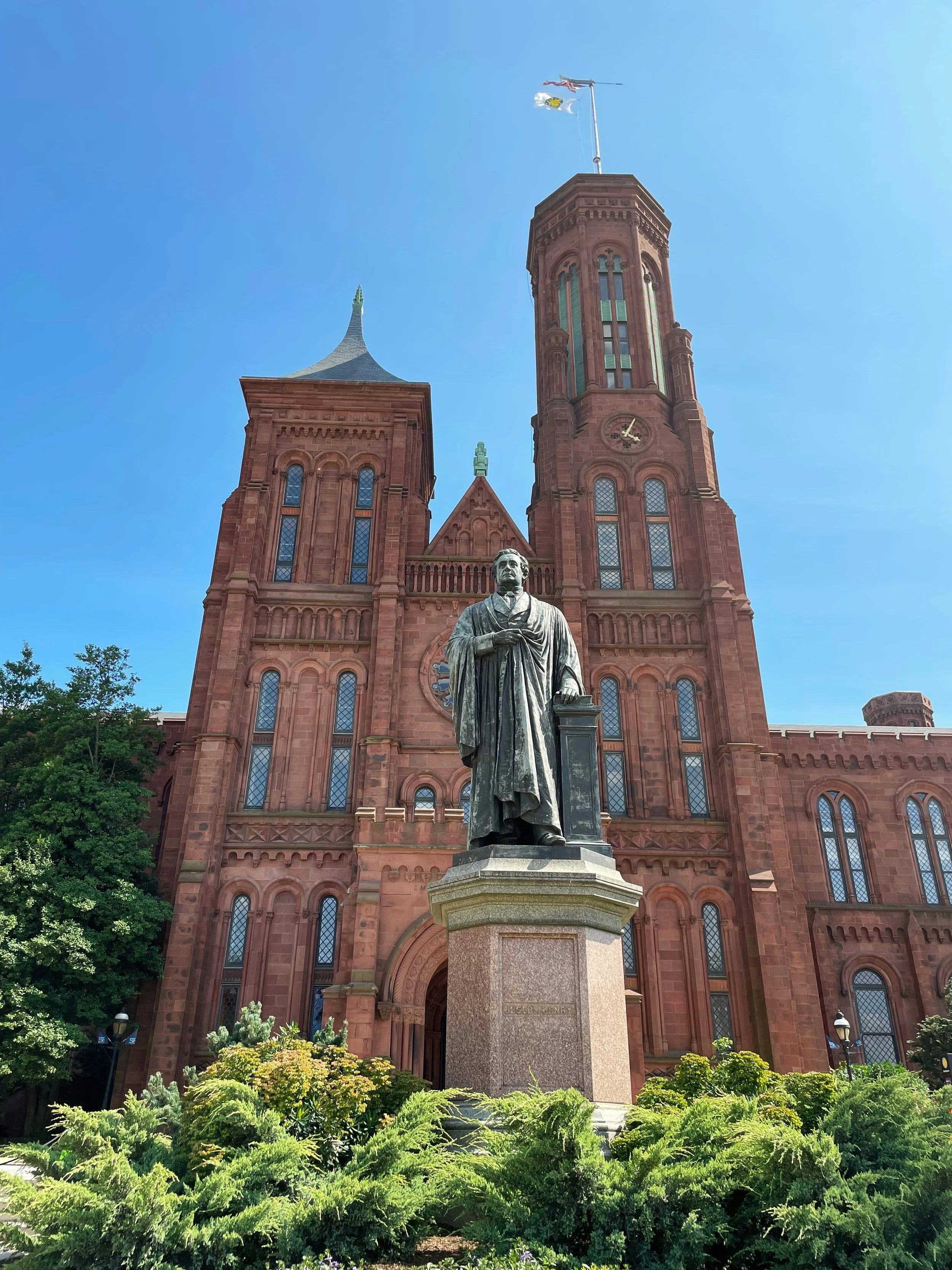 The statue of Joseph Henry stands outside the Smithsonian Castle on the National Mall in Washington DC. The statue is surrounded by green shrubs and framed by the red brick of the castle