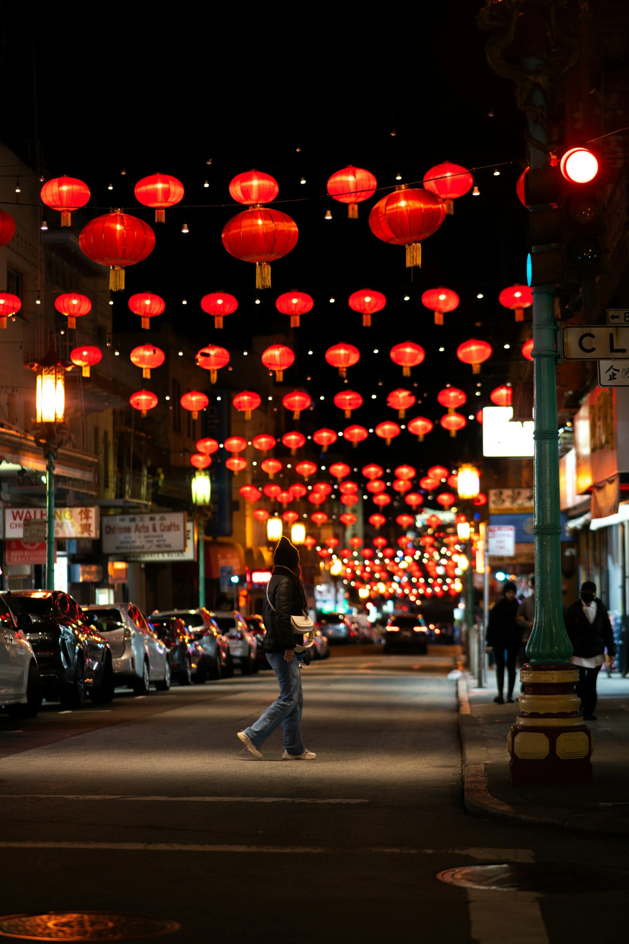 A person crosses the street in San Francisco Chinatown under lanterns that are lit up a bright orange