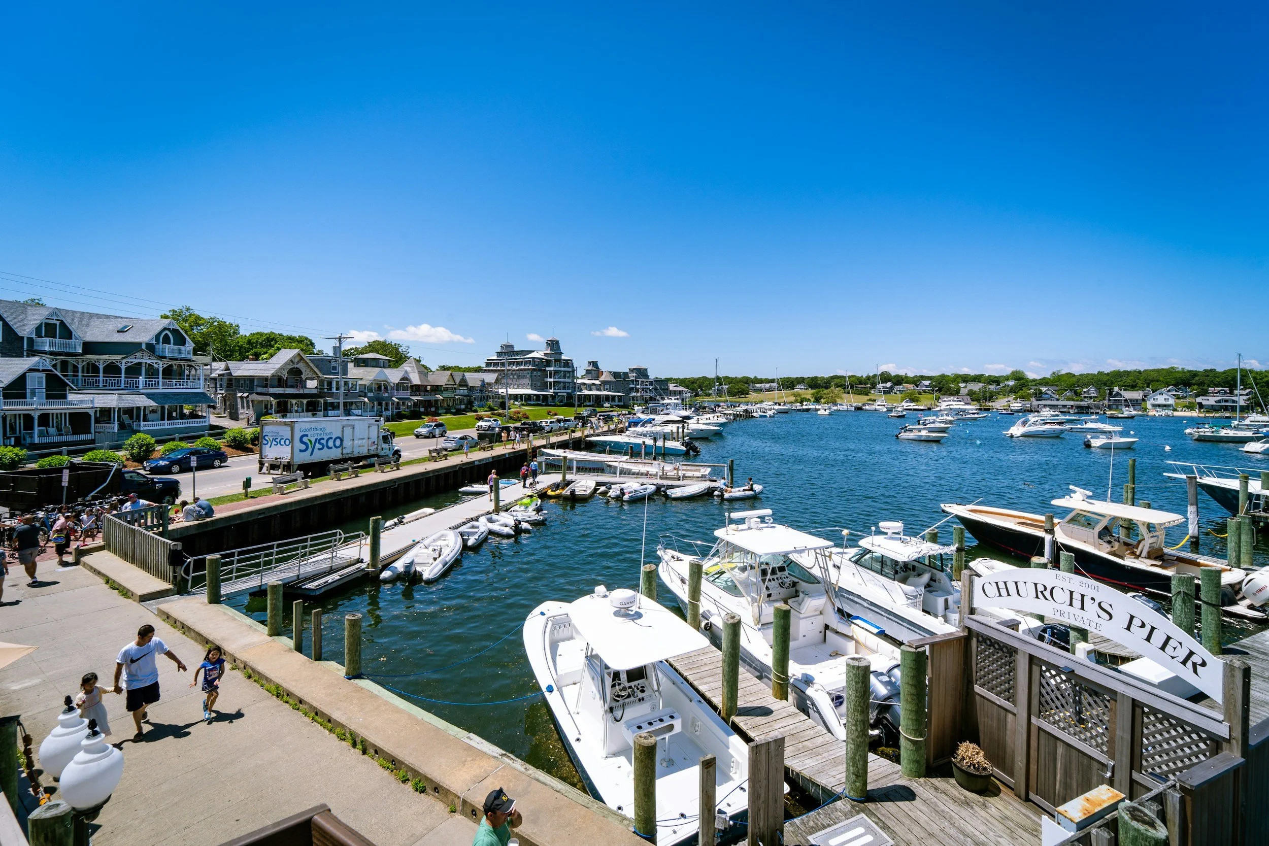 The port of Oak Bluffs on Martha's Vineyard is full of small boats. Some people walk along the waterfront and there are large houses in the distance